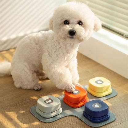 Orange Poppy pet talking button toy set with four colorful buttons on mat used by small white fluffy dog