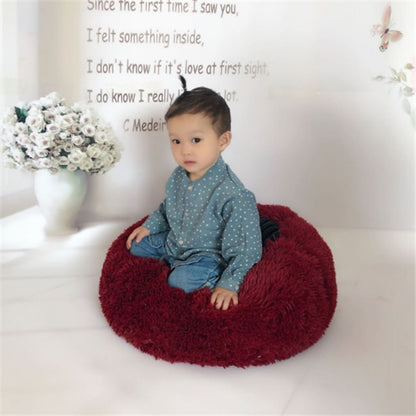 Toddler sitting on red round fluffy cat bed by Orange Poppy in bright room with floral vase backdrop