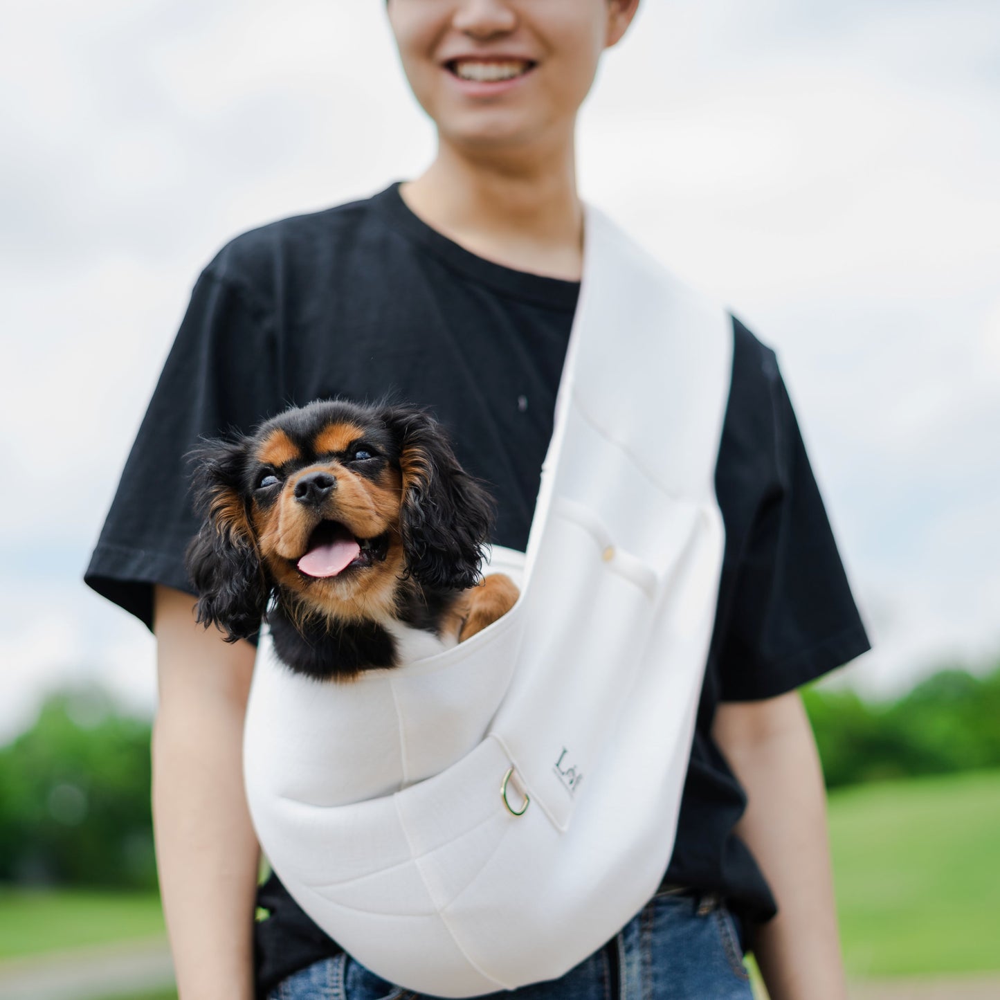 Emerald Alfie pet sling carrier for small dogs in white with a black and brown puppy inside worn by a person outdoors